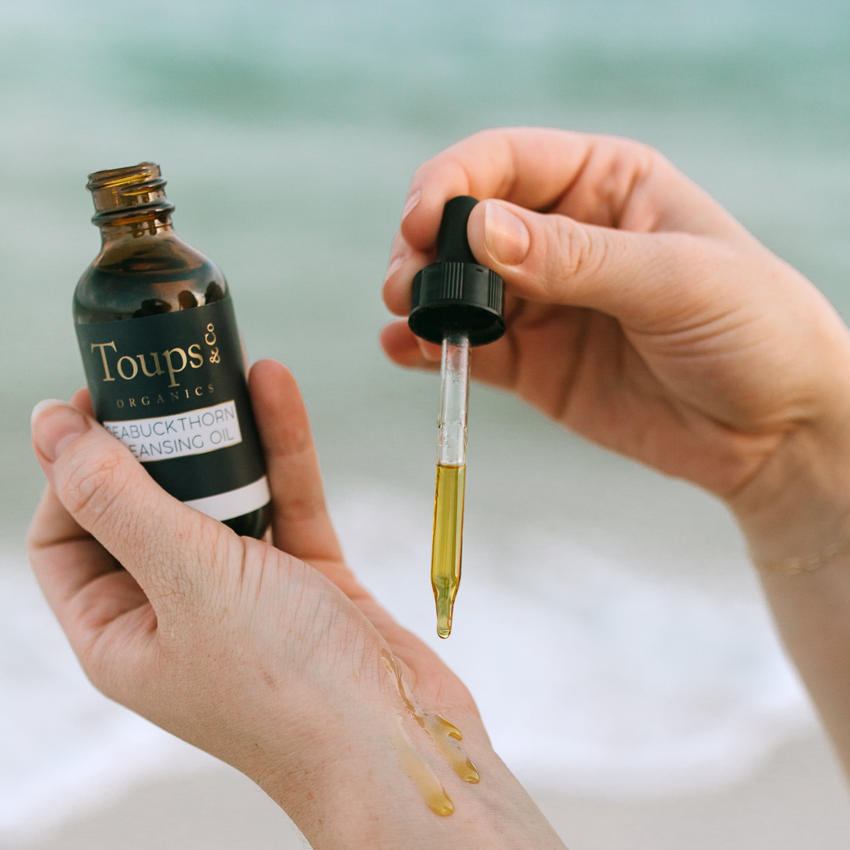 A person applies Toups and Co Organics Seabuckthorn Cleansing Oil from a dropper onto the back of their hand, ideal for sensitive skin, with a softly blurred beach background.