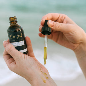 A person applies Toups and Co Organics Seabuckthorn Cleansing Oil from a dropper onto the back of their hand, ideal for sensitive skin, with a softly blurred beach background.
