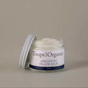 A glass jar of Toups and Co Organics Tallow Balm - Unscented (2 oz) sits with its lid off to the side, revealing the creamy balm made from grass-fed tallow inside, set against a simple beige background—a natural moisturizer ideal for sensitive skin.