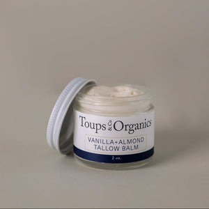 A glass jar of Toups and Co Organics Tallow Balm - Vanilla + Almond, made with nourishing grass-fed tallow, sits with the lid off to reveal the creamy balm inside, inviting you to experience ultimate skin hydration against a plain background.